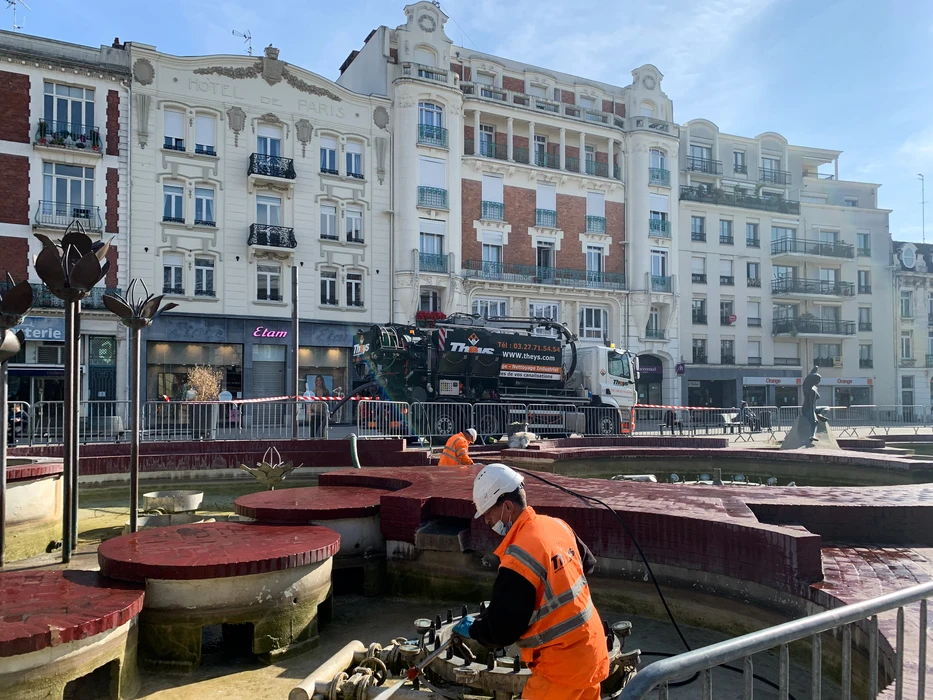 Scène urbaine en extérieur : deux techniciens en EPI (vestes haute-visibilité orange, casque blanc, masque FFP2) interviennent sur un bassin fontaine à curer