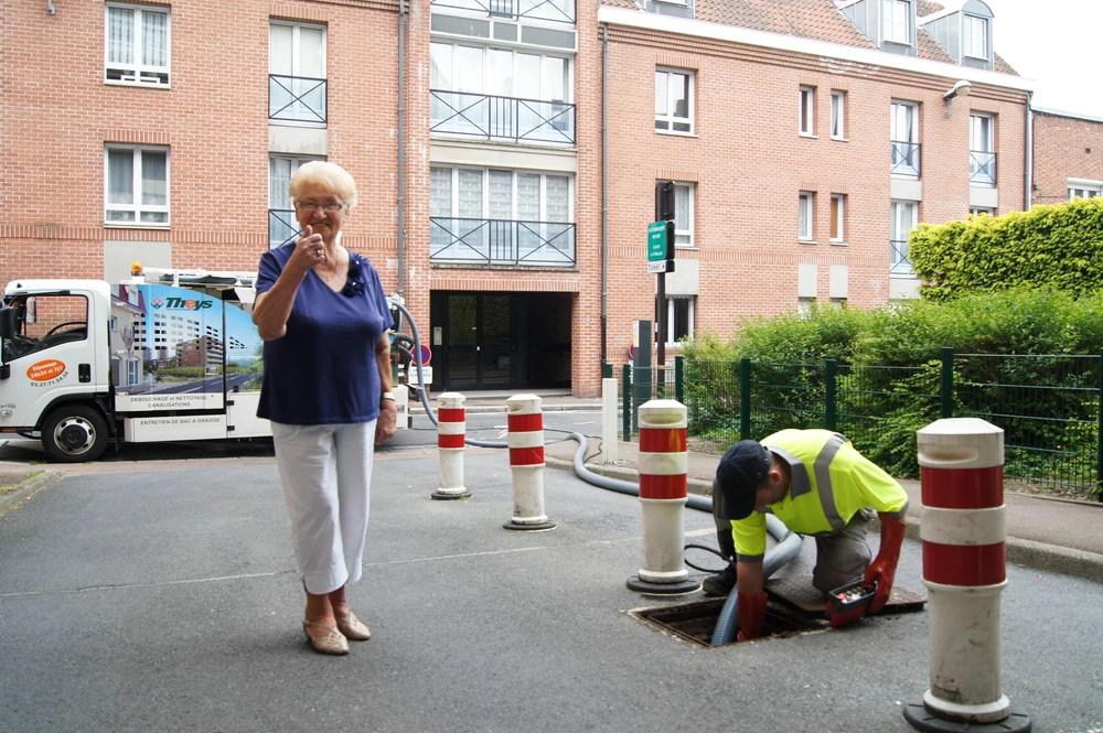Scène urbaine extérieure : une femme âgée en tenue décontractée donne un pouce levé près d'un technicien en tenue haute-visibilité intervenant sur un regard d'égout