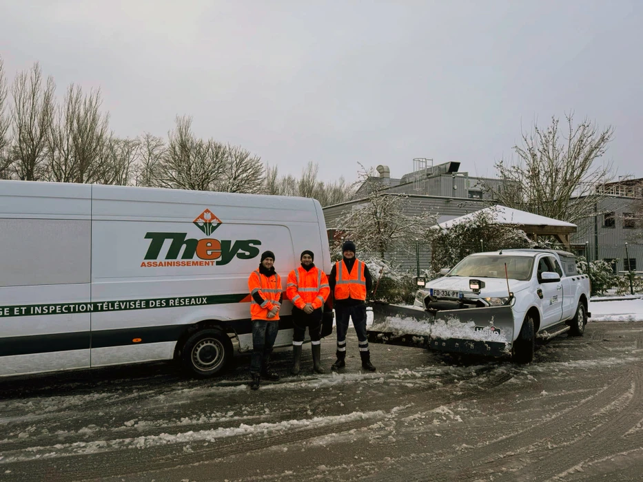 Trois techniciens en gilets haute visibilité orange debout dans un parking enneigé